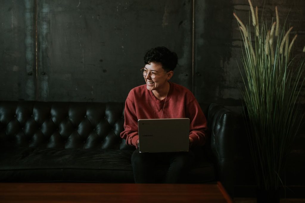 A female sits on a couch with a laptop and is smiling